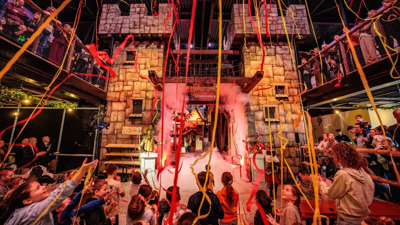 Kinderen en ouders vieren de opening van Drakendorp met slingers en rook bij een grote kasteelpoort.