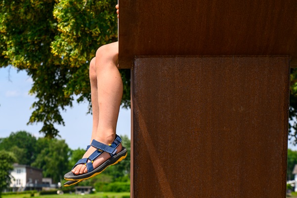 Een kind zit met bungelende benen op een groot roestbruin kunstwerk, met op de achtergrond bomen en een huis aan de Maas.