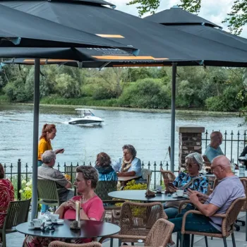 Gasten zitten op het terras van Brasserie LUS in Linne, met uitzicht over de Maas en een boot op het water.